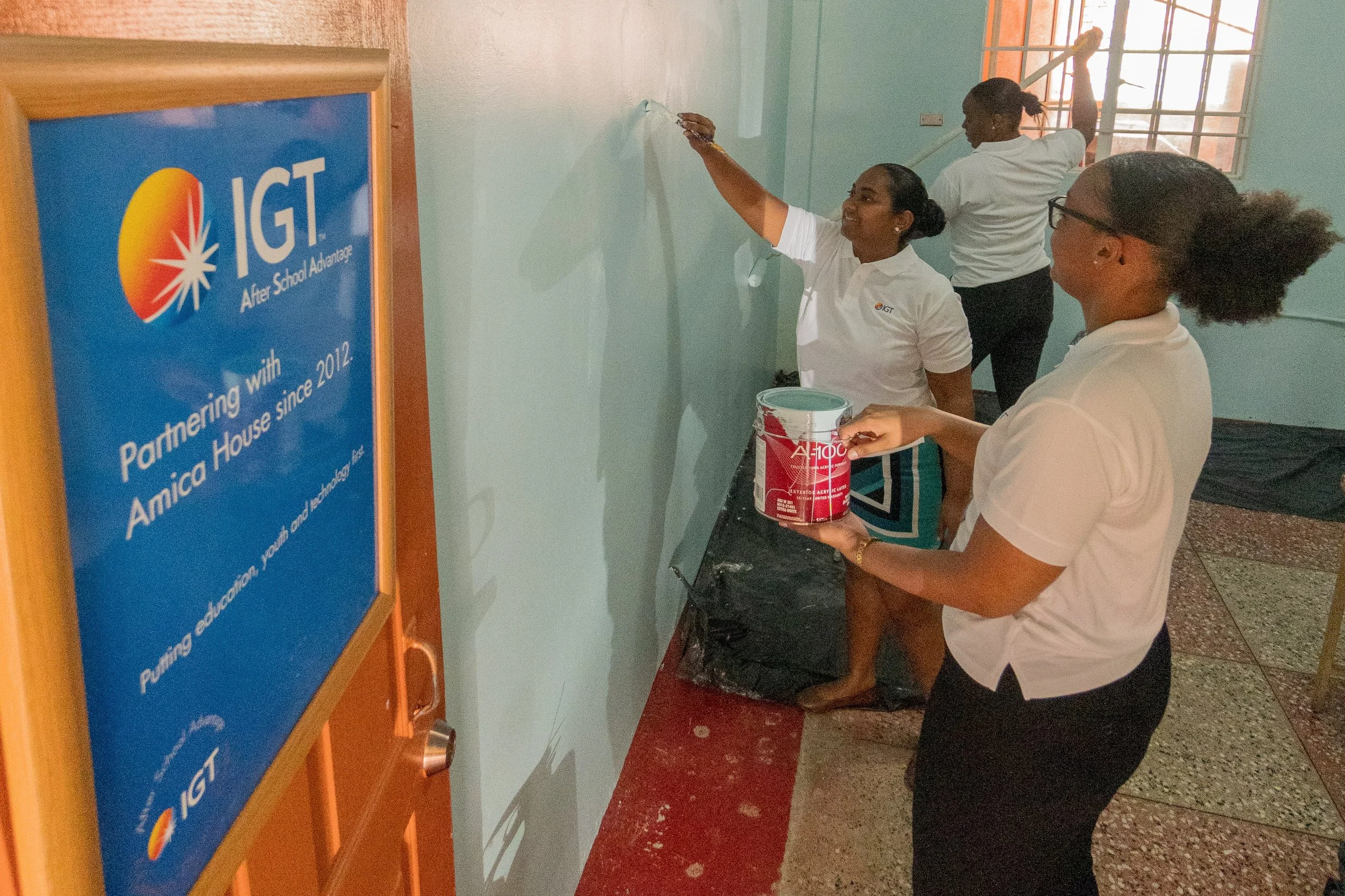 Hard at Work! IGT’s volunteer team members, Tanya Singh, IGT Global People Services Representative (centre), Akilah Howard, IGT Call Center Associate (back), and Chelsea Alleyne, IGT Administrative Assistant (front), show off their painting skills during their Global Giving Month volunteer work at Amica House children’s home in May 2024.